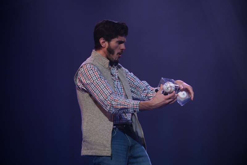 Man in plaid shirt and vest holding two baseballs in clear cases, mouth open, against a dark blue background.