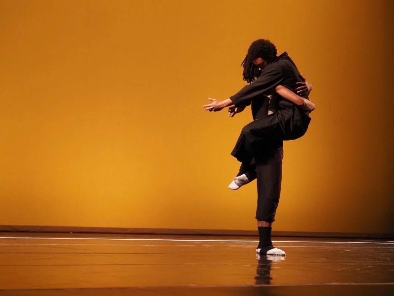 Performance art on a stage with a yellow backdrop. Two figures, one lifting the other in an embrace. Both are dressed in black, with white socks visible on the lifted figure. Stage floor reflects the warm light.