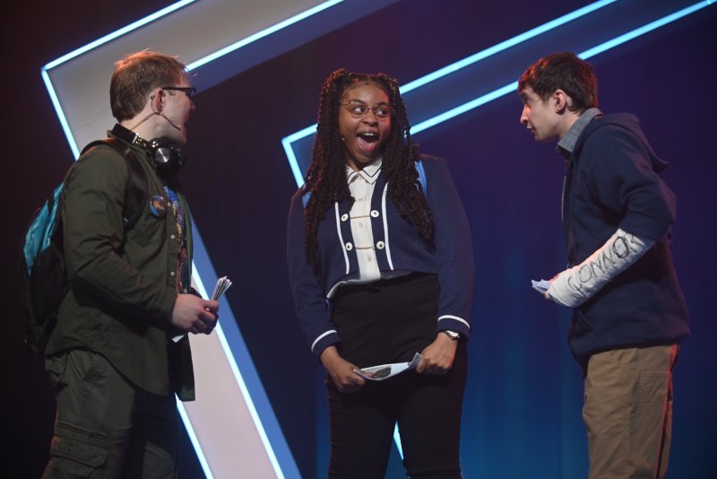 Three young adults on stage, lit by blue and purple lights. A Black woman in the center looks surprised, flanked by two white men.