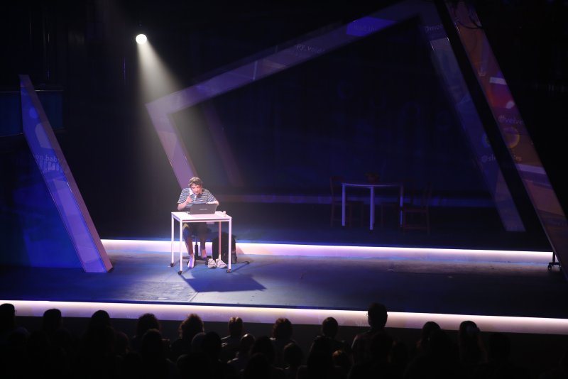 A lone actor sits at a white desk with a laptop, illuminated by a spotlight on a dark stage with glowing blue geometric screens.