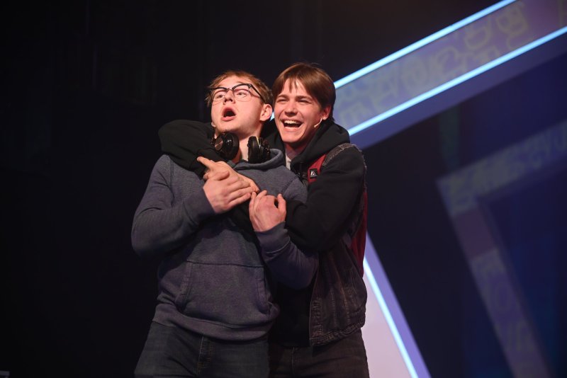 Two young men on stage, one with glasses looking up, the other behind him laughing, arms around his neck.