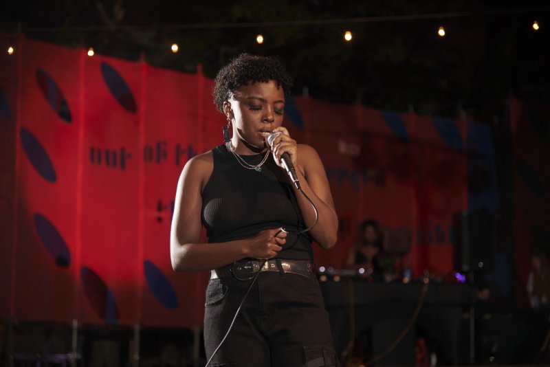 A performer sings into a microphone. She wears a black tank top and shorts, with jewelry. A red backdrop with blue oval shapes and text is behind her. String lights are visible above.