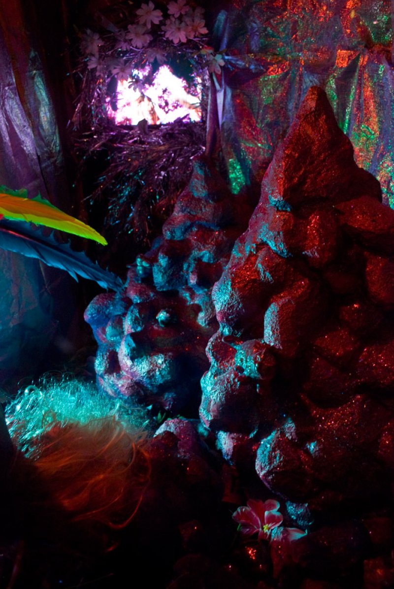 Abstract installation with textured, rock-like forms in red and blue hues. Shimmering fabric backdrop, floral accents, and a brightly lit opening at the top.