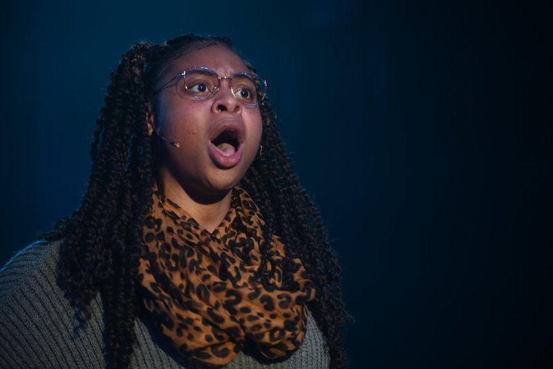 A young woman with braided hair and glasses, wearing a leopard print scarf, sings with her mouth open against a dark background.