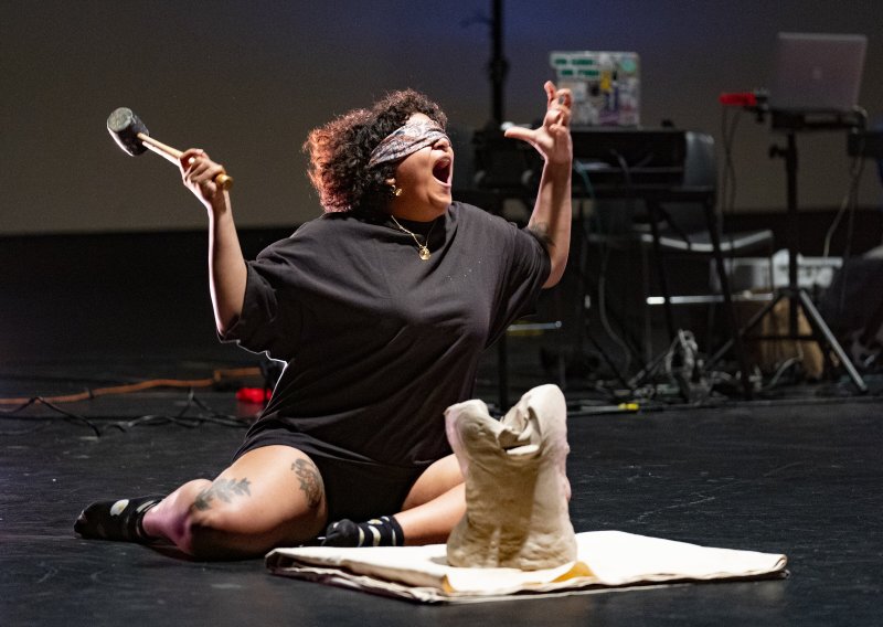 Blindfolded performer sits on stage, mouth open, holding hammer aloft. A clay sculpture rests on a cloth in front of them. Dark clothing contrasts with the bright stage light.