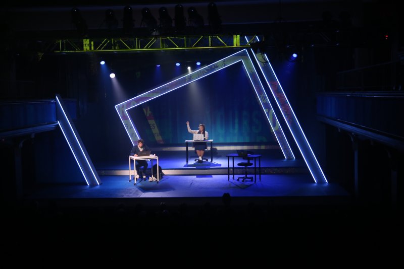 Two actors on a dark stage with glowing blue neon frames, one seated at a desk, the other standing with a laptop.