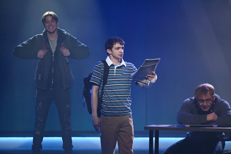 Three men on a stage with blue lighting. One man in a striped shirt holds paper, another in a dark jacket smiles, and a third man in glasses sits at a desk.