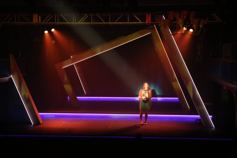A woman stands center stage, illuminated by spotlights, amidst a modern set of glowing geometric frames under warm and cool lighting.