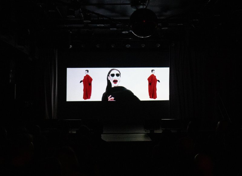 Video art with a central figure in black screaming, flanked by two figures in red robes, all on a bright screen in a dark room.