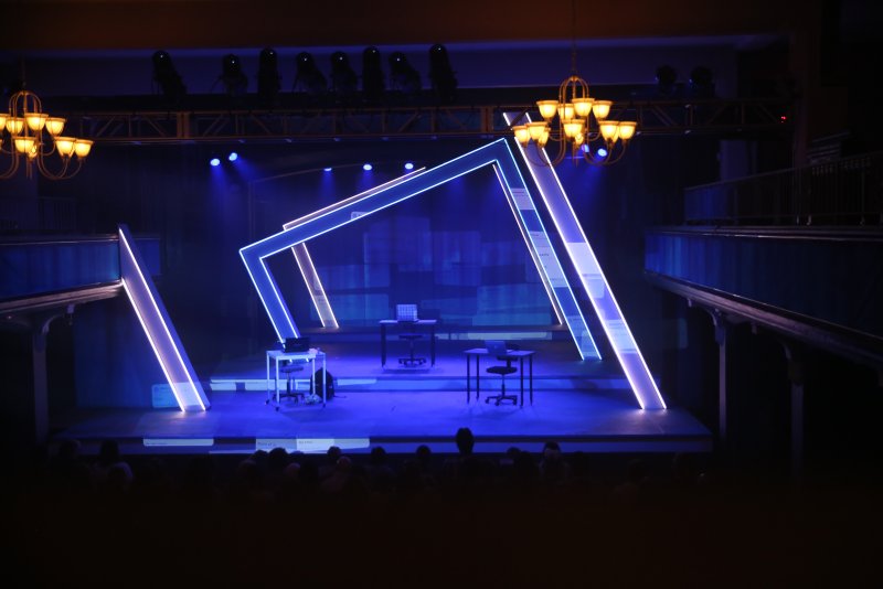 Stage with blue lighting, three desks, and glowing white geometric frames, viewed from audience seating.