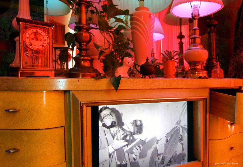 Wood cabinet with open drawer revealing a black and white video of a man on the phone. Cabinet top cluttered with lamps, plants, clock and figurines, bathed in pink and blue light.