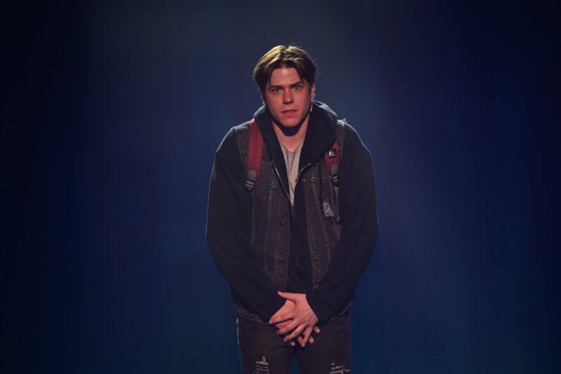 A young man with brown hair, wearing a dark hoodie, denim vest, and red backpack, stands with hands clasped, looking forward.