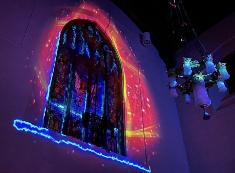 Stained glass window illuminated by a vibrant, swirling projection of red, pink, and blue light, with a blue neon outline at the bottom.