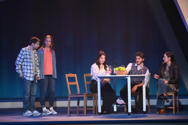 Four actors on a stage with a dark blue background. Two stand left, two sit right at a white table with apples and wine.