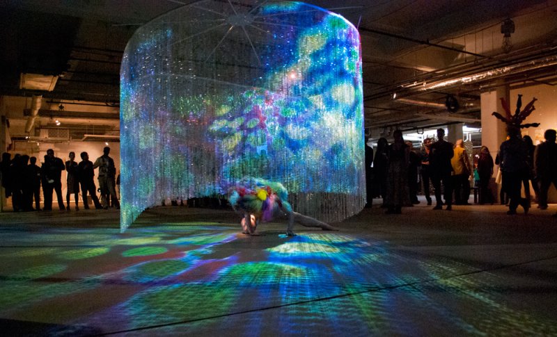 Performance art with a dancer in a colorful costume under a shimmering, iridescent curtain, casting vibrant light patterns.