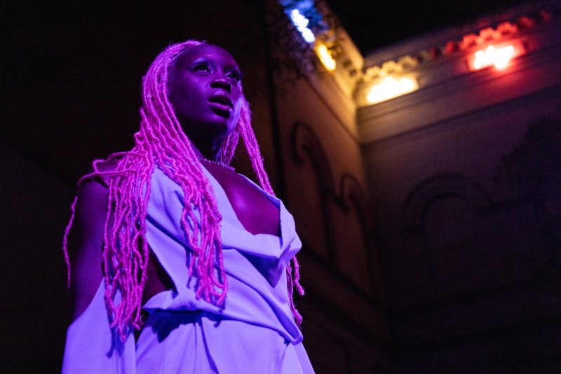 A Black person with long pink dreadlocks, wearing a light blue garment, looks up and to the right under purple and blue stage lighting.
