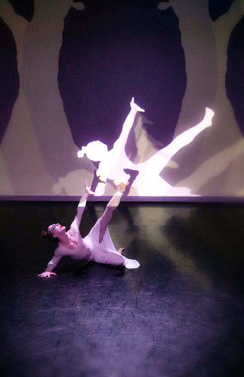 Dancer in white dress on dark stage, limbs extended, lit by purple light. Behind, a large projected silhouette mirrors the pose, framed by abstract shapes.