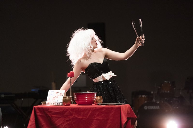 Performance art scene: a person with white curly hair and a black sequined dress stands behind a red table, holding tongs up to their face. A red candle and bowl sit on the table. Dark background.