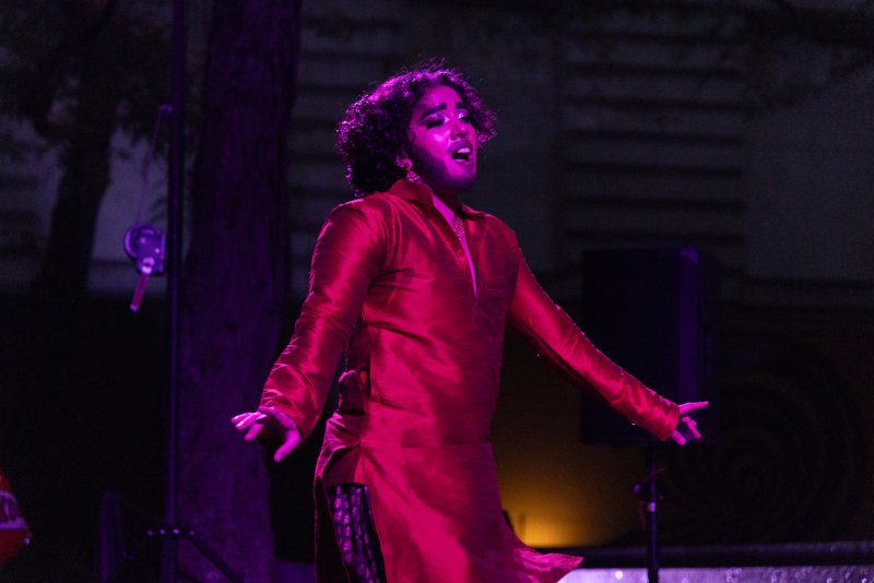 Person in red silk tunic with open arms, lit with purple light, singing. Curly hair, beard, and glittery makeup. Dark background.