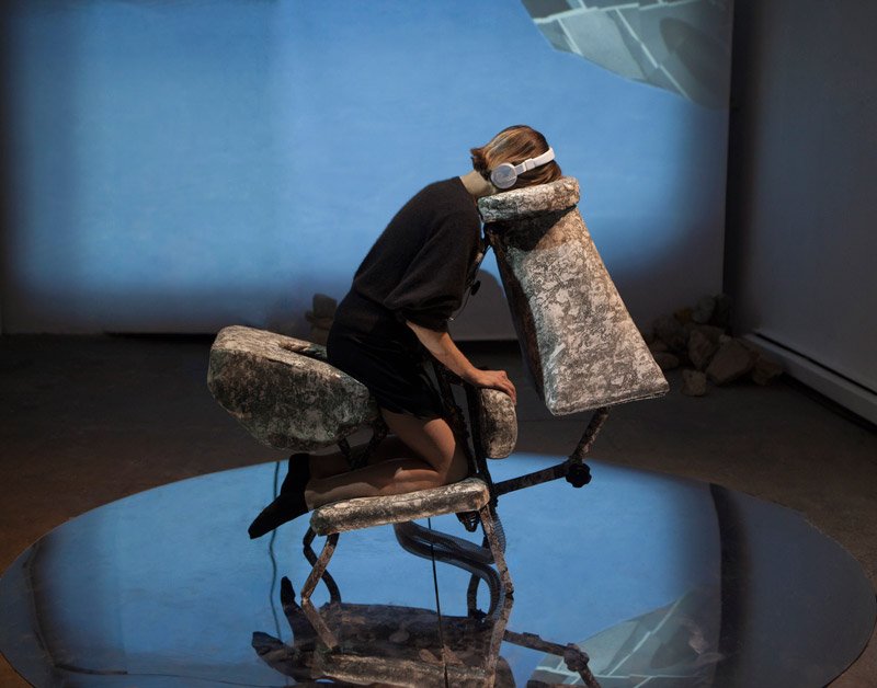 Woman in headphones and black clothing hunched over a textured, grey, abstract sculpture, reflected on a circular mirror.