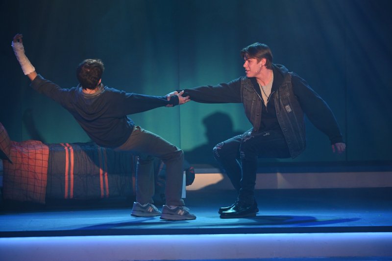 Two young men on stage, one with a cast, holding hands, bathed in blue light.