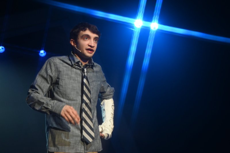A young man in a striped tie and arm cast sings on stage under bright blue spotlights.