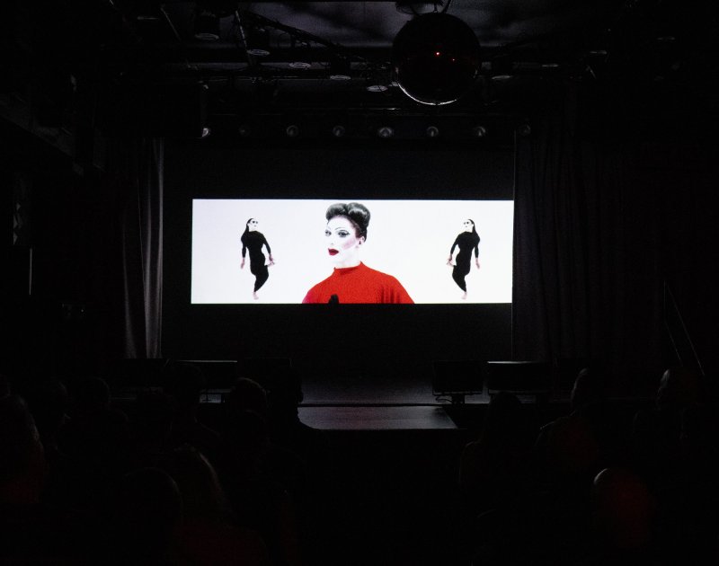 Drag queen in red top with two dark figures on white screen, viewed by silhouetted audience in dark room.