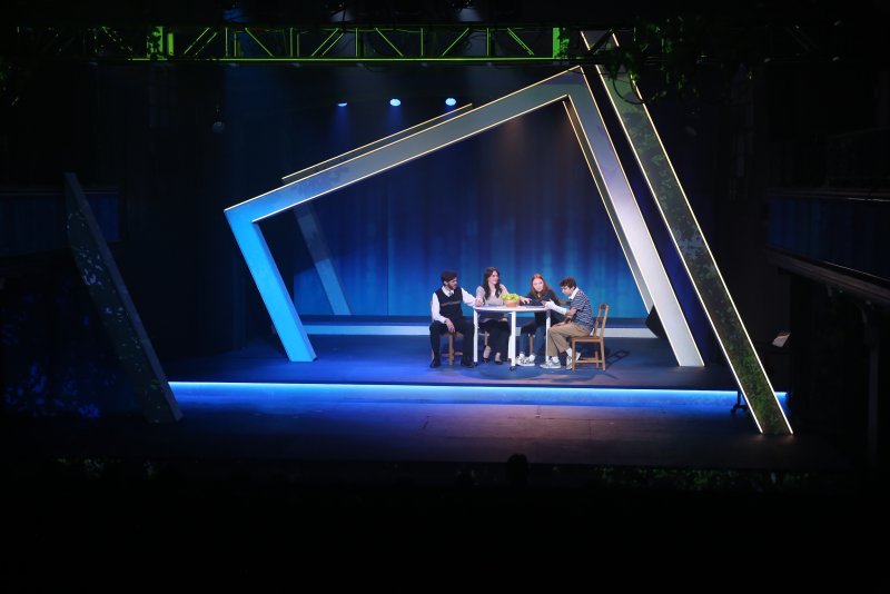 Stage set with four actors at a table, framed by an illuminated geometric arch against a dark blue backdrop.