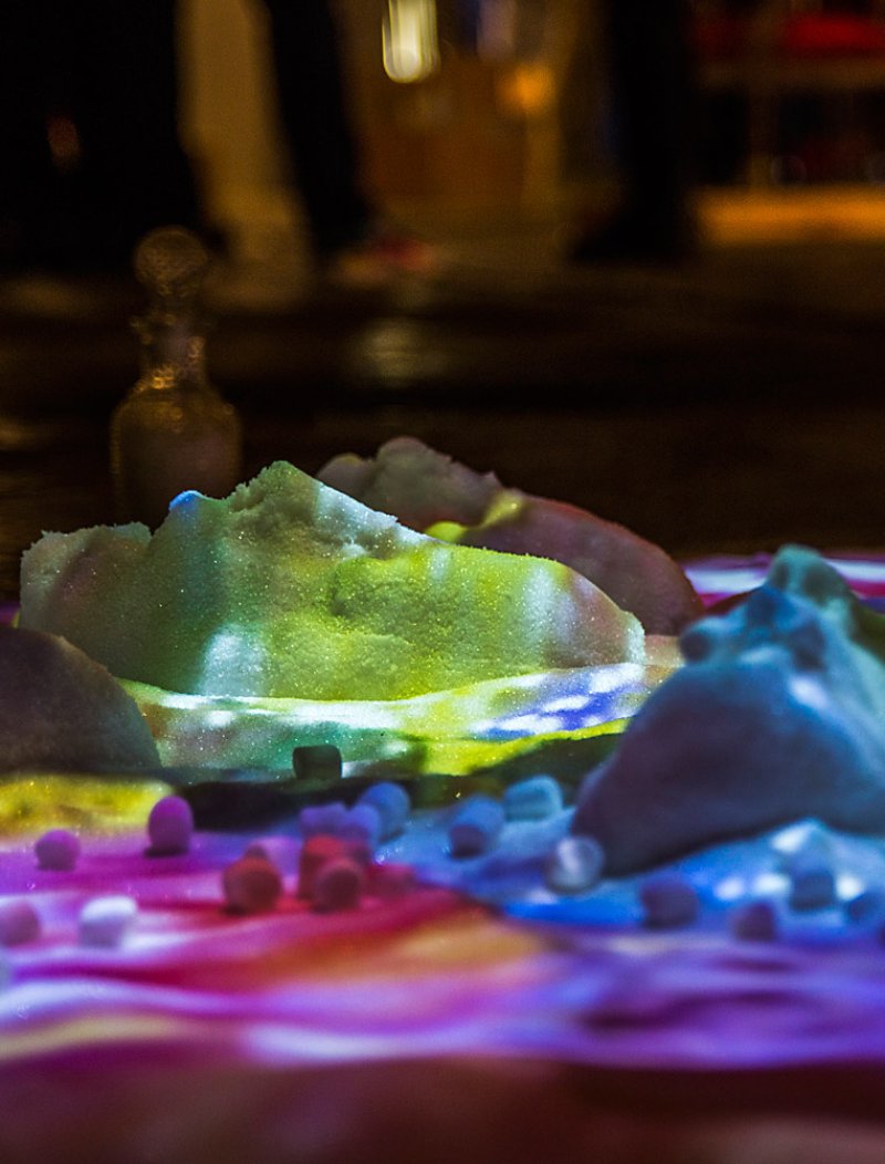 Mounds of sugar illuminated by projected colorful lights, with small candies scattered across a vibrant surface.
