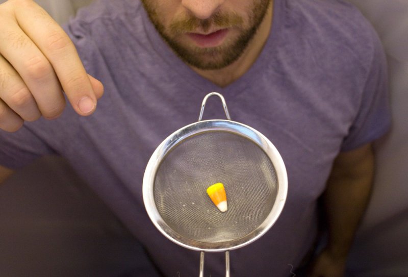 A man in a purple shirt holding a single candy corn in a metal strainer, looking down at it.